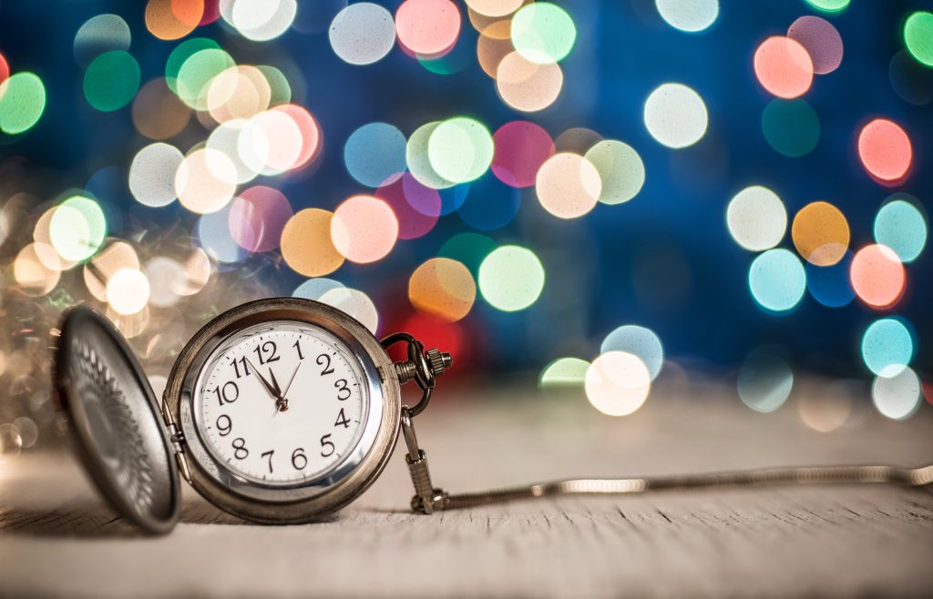 A pocket watch clock just before midnight with colorful lights in the background.